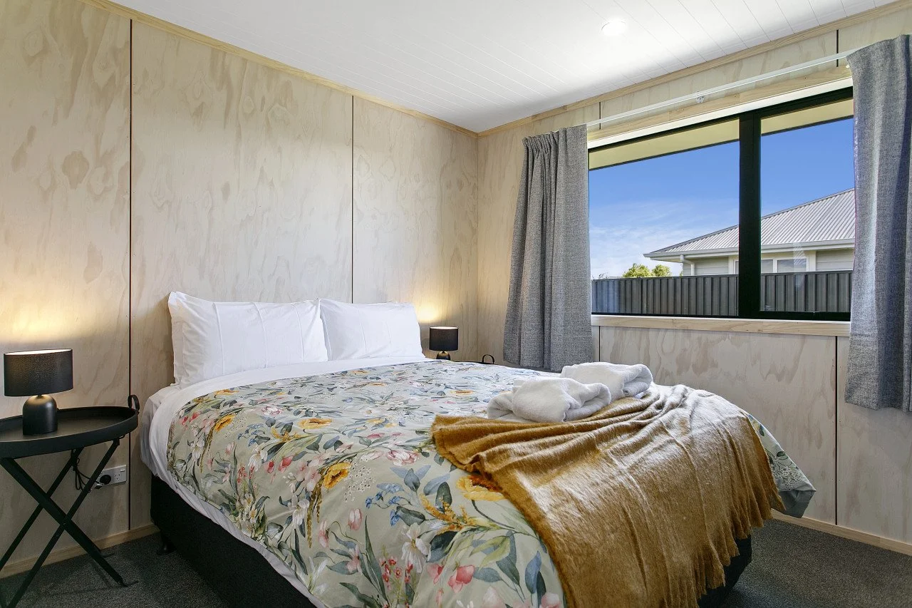 A bedroom with a bed, floral bedding, folded white towels, a brown blanket, two bedside tables with black lamps, a large window with gray curtains, and a view of neighboring houses and blue sky.