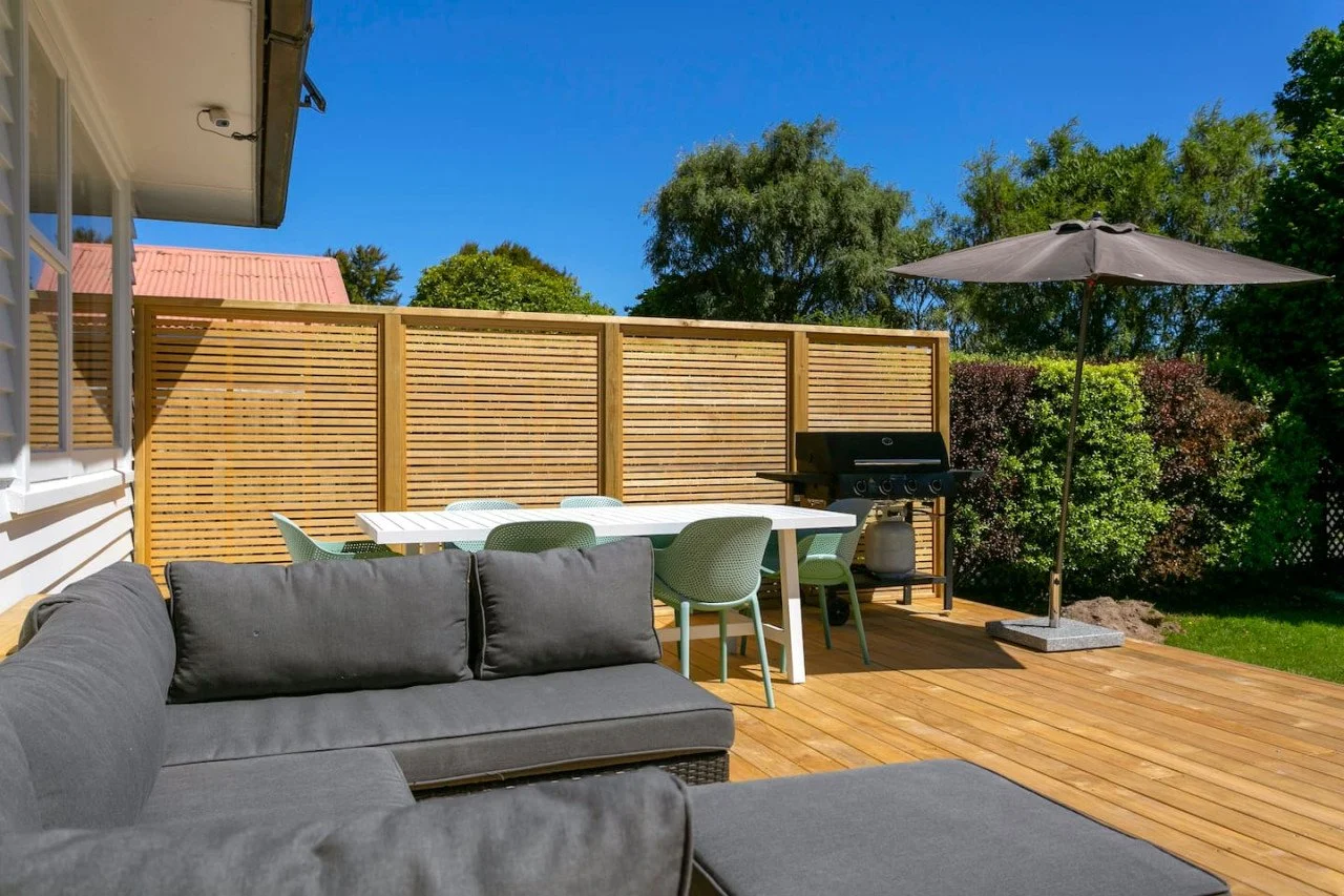 Outdoor backyard patio with gray sectional sofa, white dining table with green chairs, black barbecue grill, large umbrella, wooden deck, privacy fence, green trees, blue sky.