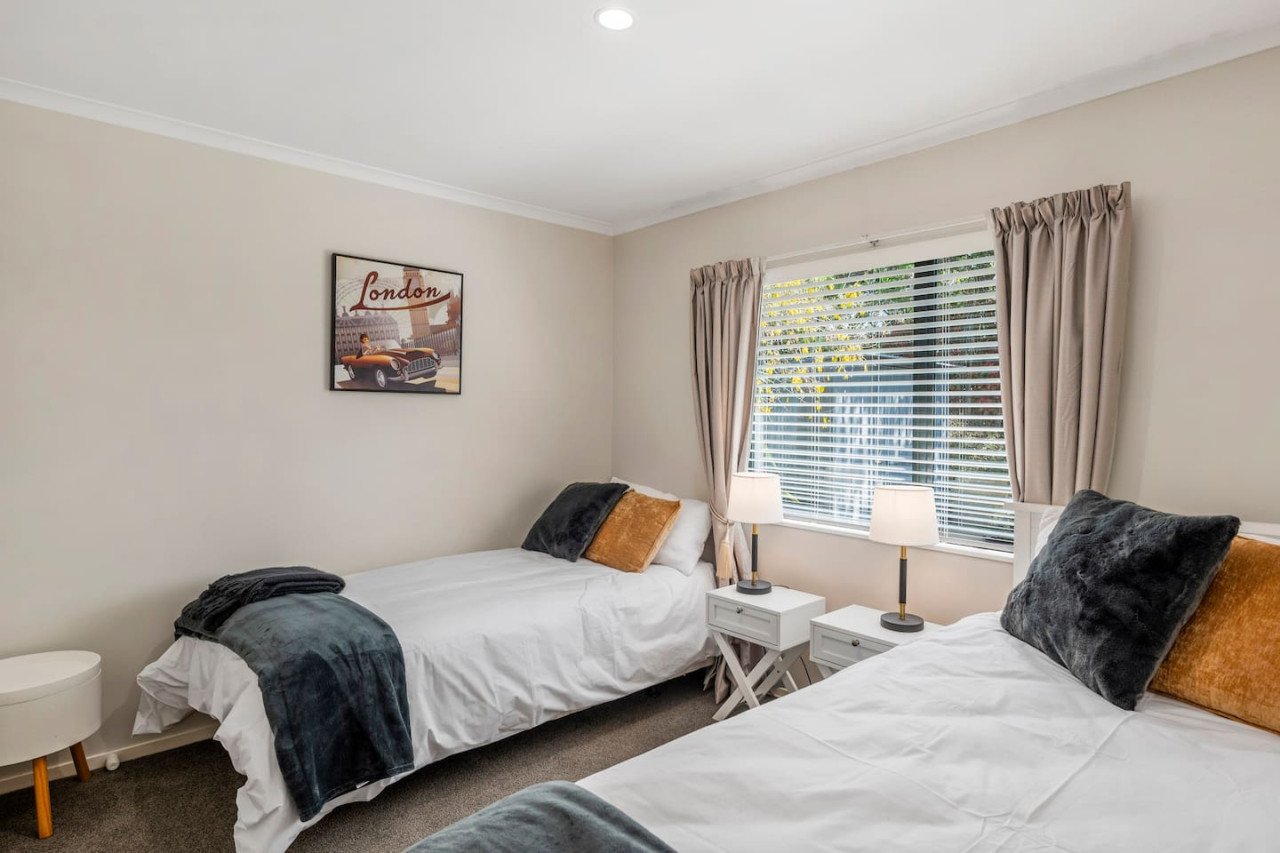 A bedroom with two beds, each with dark and beige pillows, white bedding, and a dark throw blanket. There's a window with blinds and beige curtains, flanked by two white nightstands with lamps. A framed picture of a vintage car and the word 'London' 