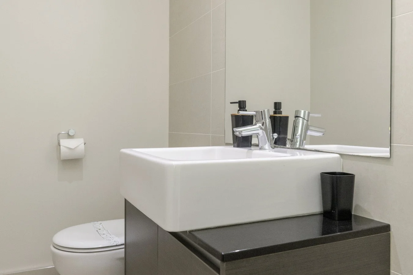 Bathroom sink with a mirror, soap dispensers, a cup, and a toilet in a minimalist setting.