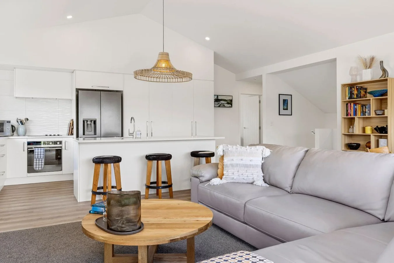 Modern open-plan living room and kitchen with white cabinets, gray sofa, wooden coffee table, and bar stools.