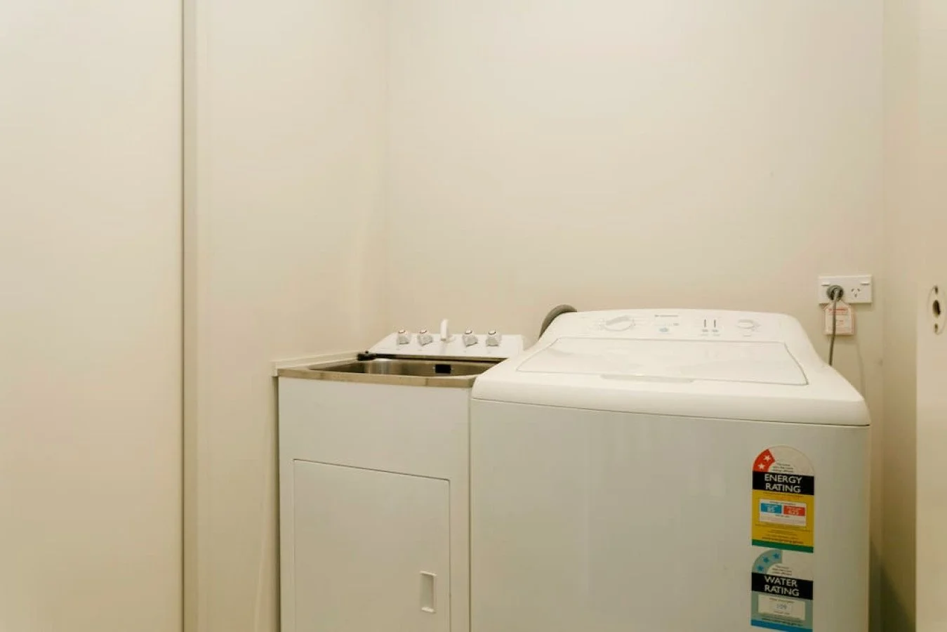 A small laundry room with a washing machine on the right and a small utility sink on the left, all against plain beige walls.