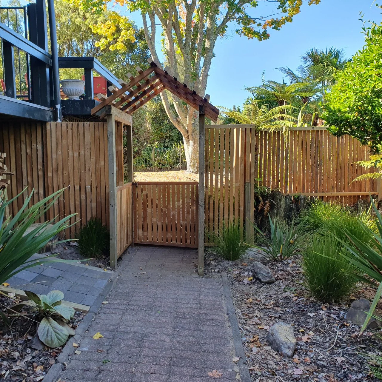 backyard garden with a wooden gate and fence, surrounded by plants, trees, and rocks under a clear blue sky.