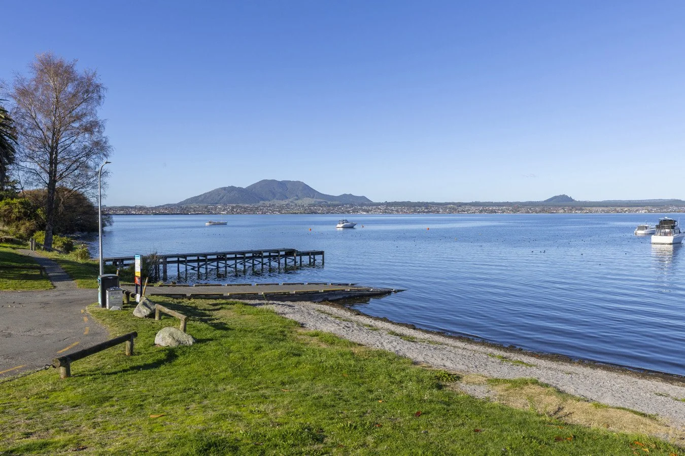 A peaceful lakeside scene with a grassy shoreline, boats on the water, and mountains in the distance under a clear blue sky.