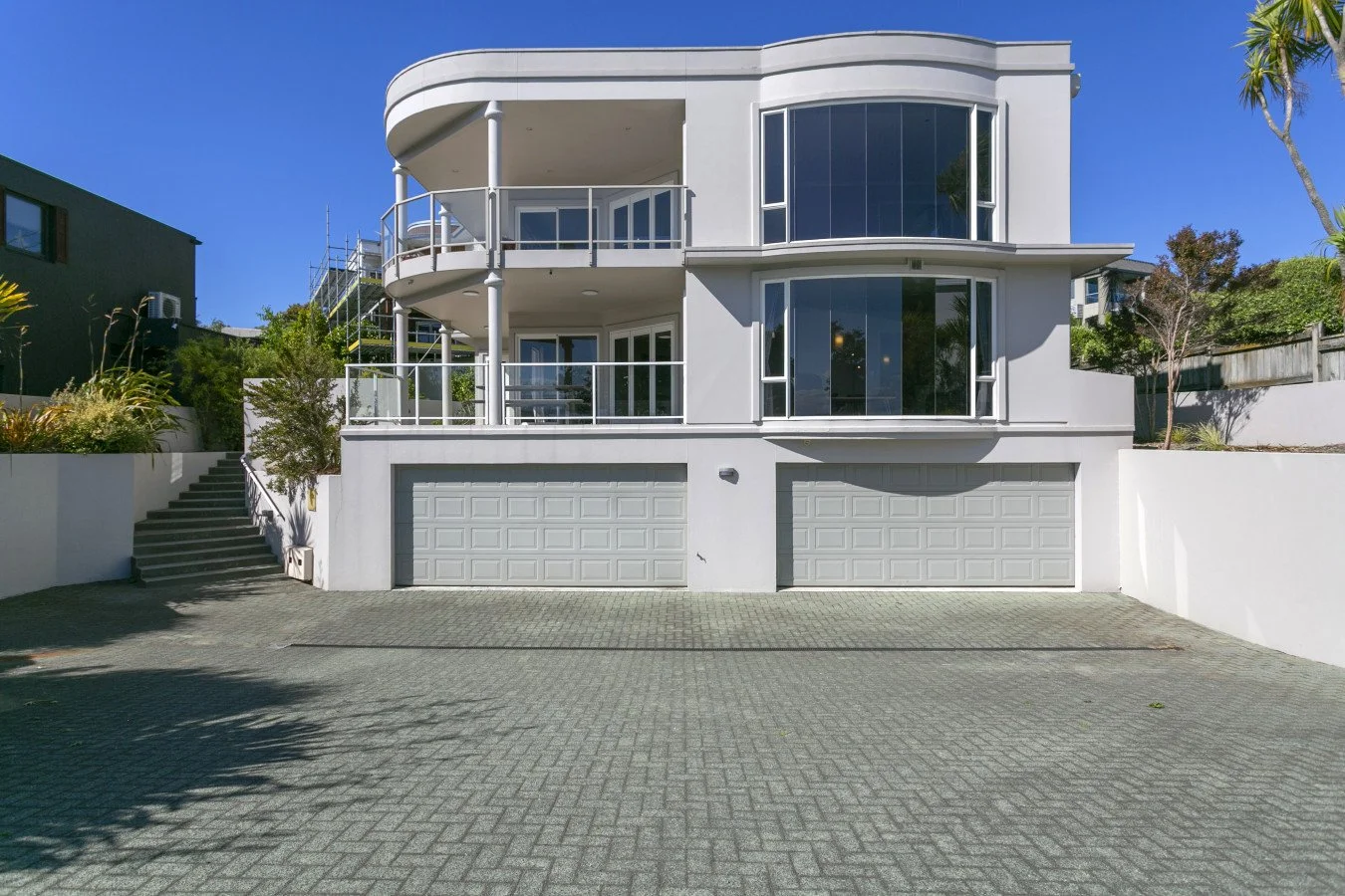 Modern white multi-story house with curved large windows, balconies with railing, and two garage doors, surrounded by trees and greenery in a sunny setting.