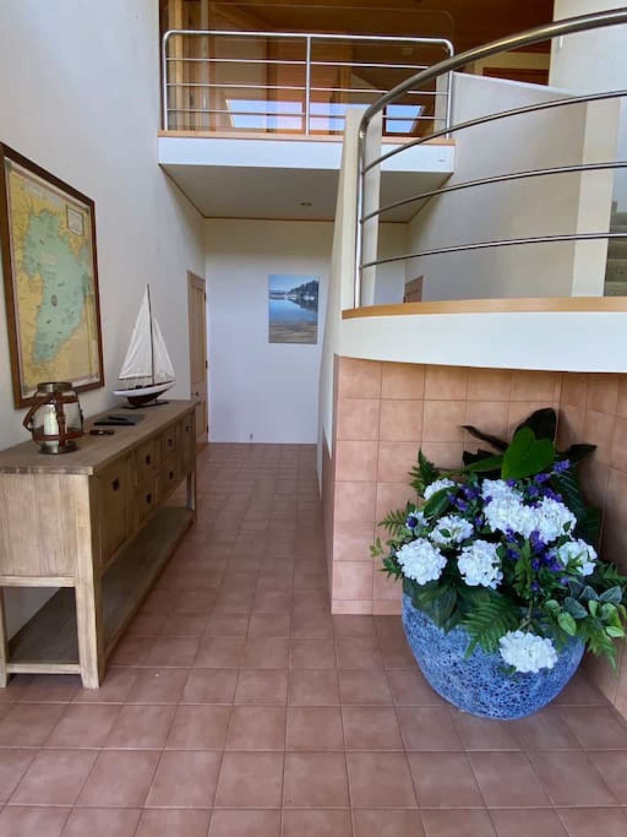 Interior view of a house with tile flooring, a wooden sideboard, ocean-themed artwork, a model sailboat, a large floral arrangement in a blue pot, and a staircase leading to a balcony with metal railings.
