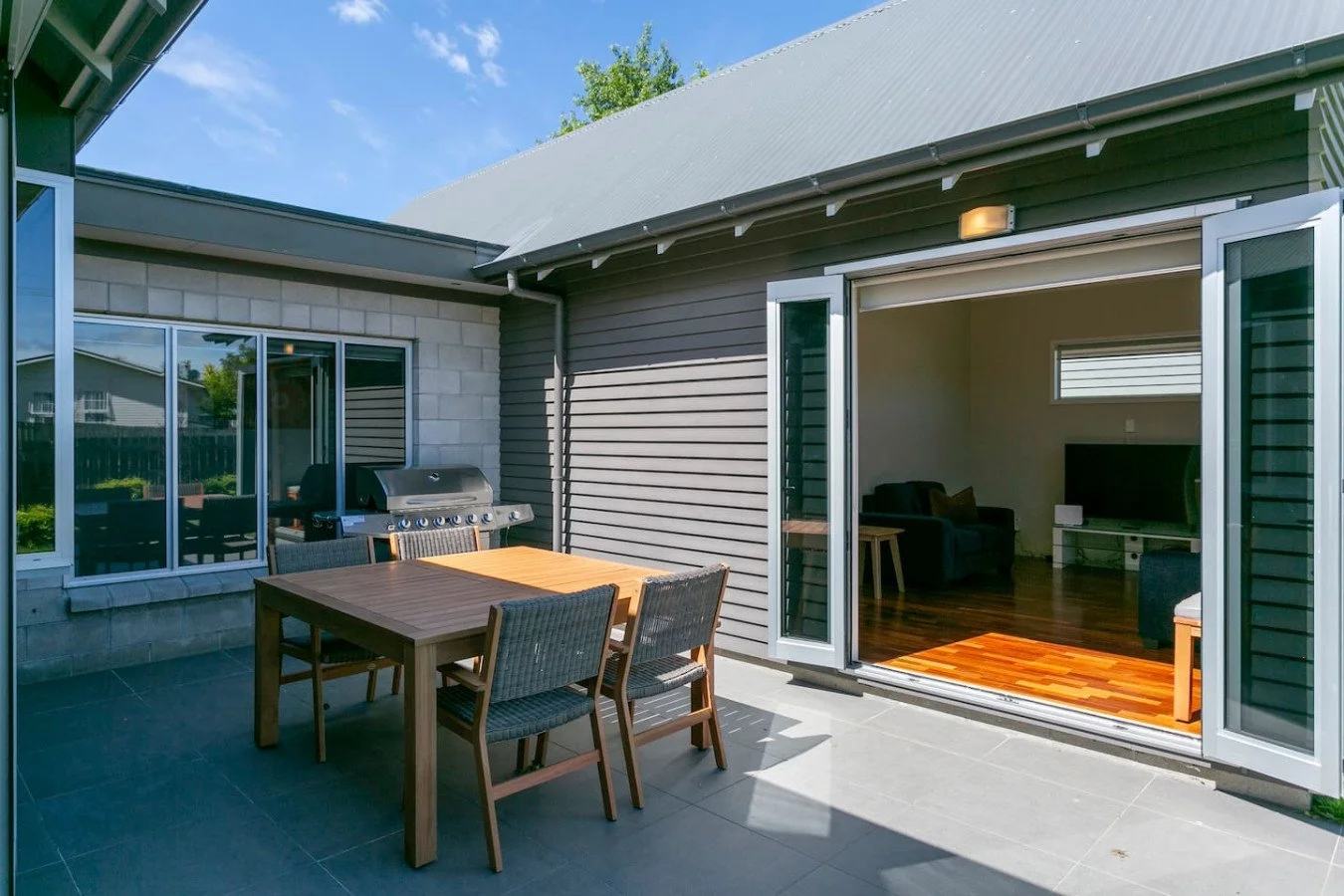 Outdoor patio with a wooden dining table, six chairs, a gas grill, and sliding glass doors leading into a living room with hardwood floors, a black sofa, and a positive bright ambiance.