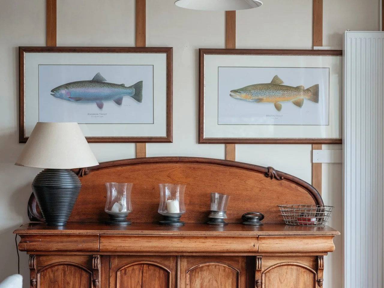 A wooden sideboard with various decor items including candles and a lamp, above which are two framed fish illustrations on a wall.
