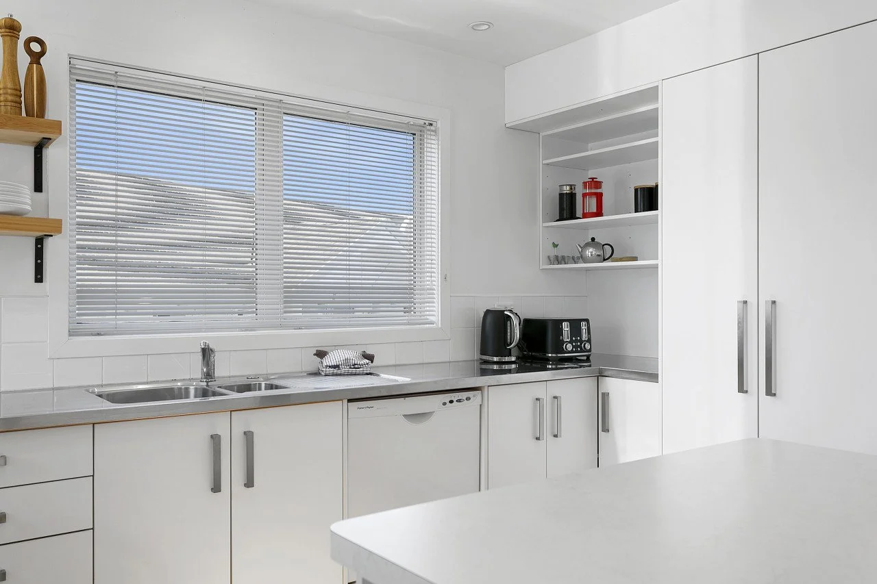 Modern white kitchen with window blinds, countertops, and appliances including a kettle and toaster.