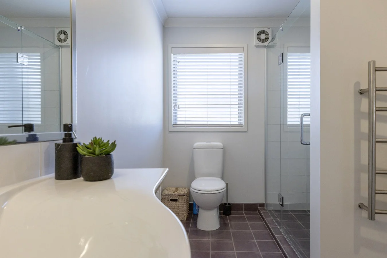 Modern bathroom with white walls, a window with blinds, a toilet, a glass shower with a door, and a white countertop with small plants.