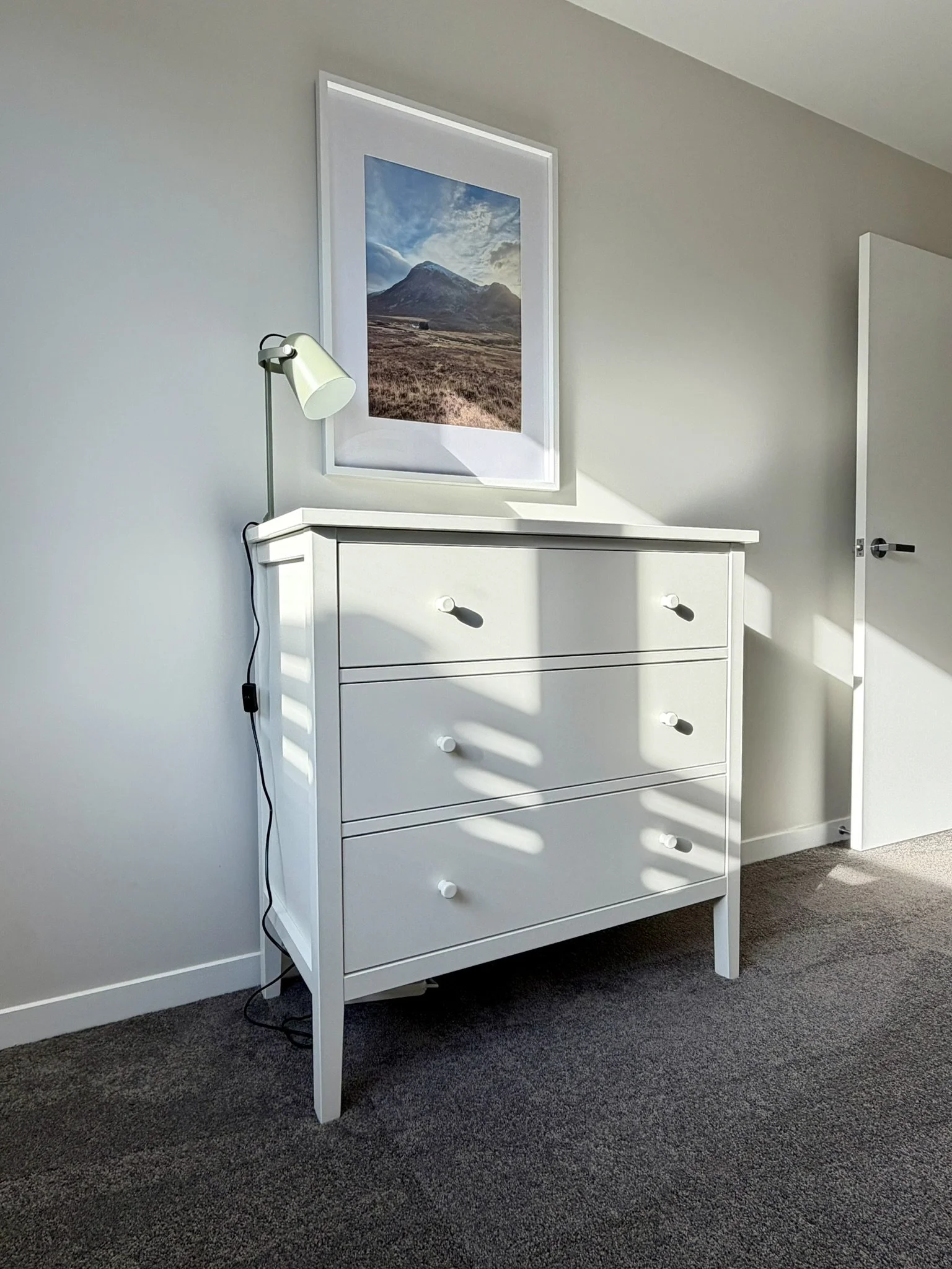 A white dresser with three drawers and round knobs, standing against a wall with sunlight casting shadows, above which hangs a framed picture of a mountain landscape. A green table lamp is leaning on the dresser, plugged into an outlet.