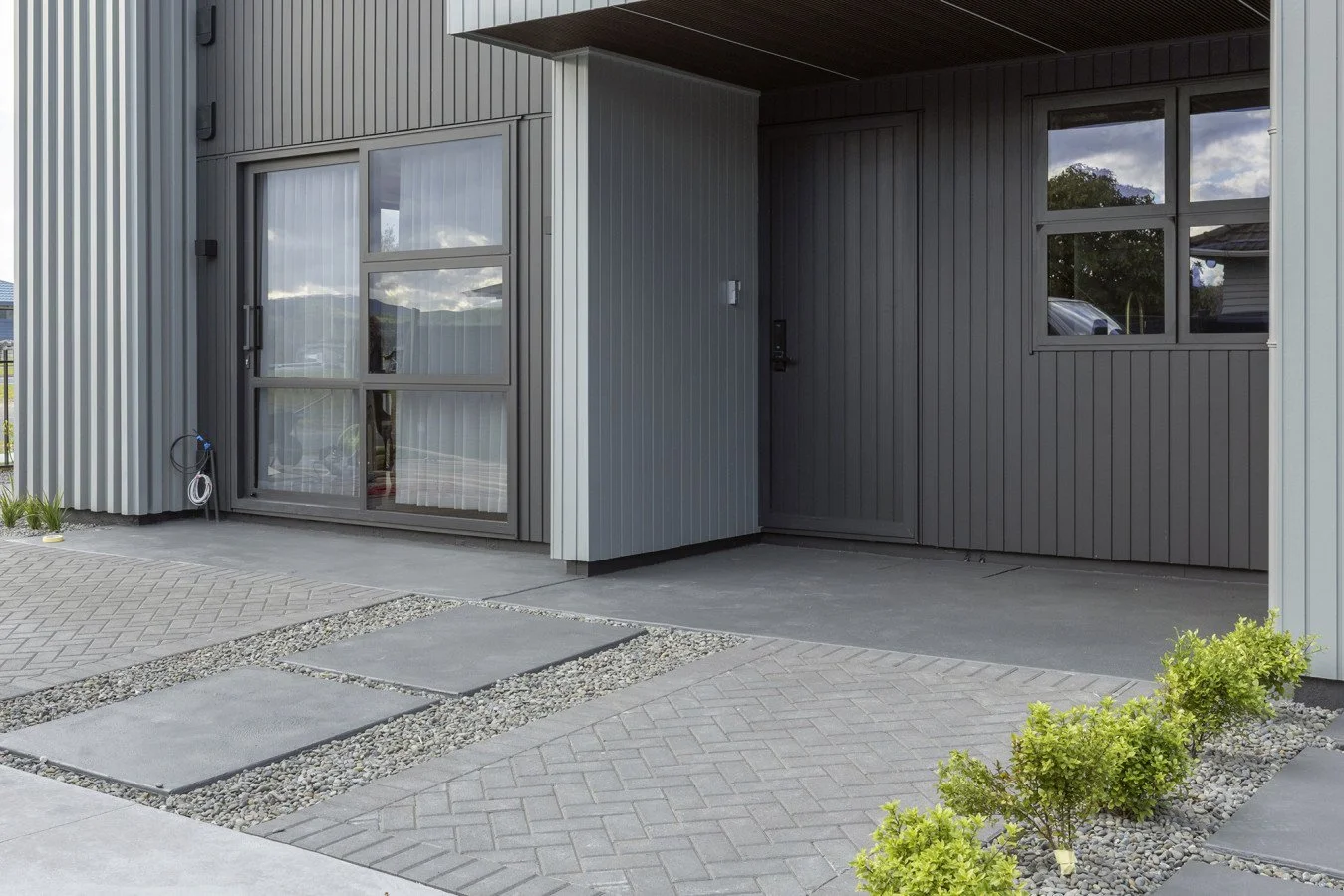 Modern house exterior with black and grey vertical metal siding, concrete walkway with large pavers and small landscaping plants.