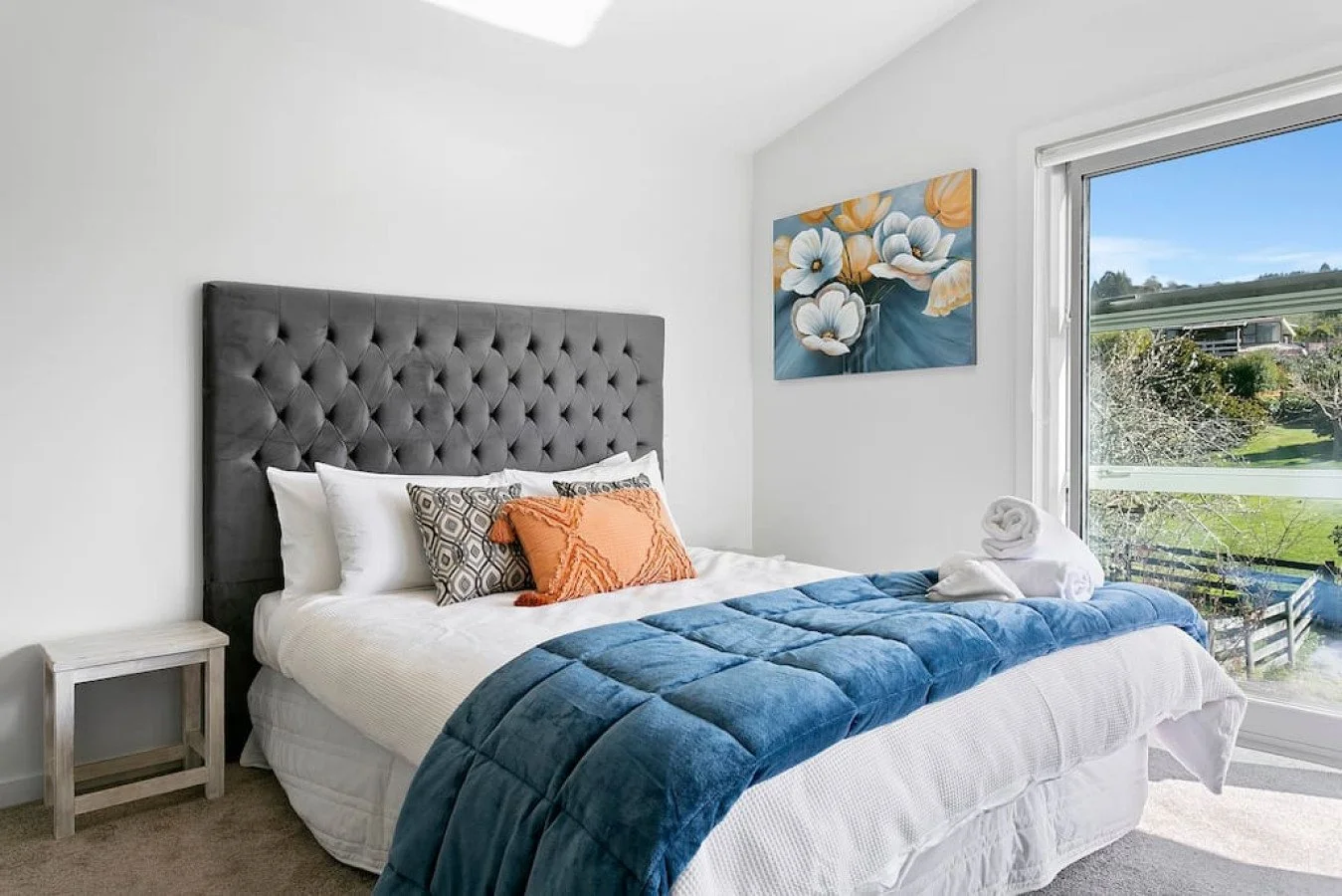 Bright bedroom with a large bed featuring a dark gray tufted headboard, white bedding, assorted pillows, a folded blue blanket at the foot, a small white nightstand, and a large window showing a lush green outdoor landscape.