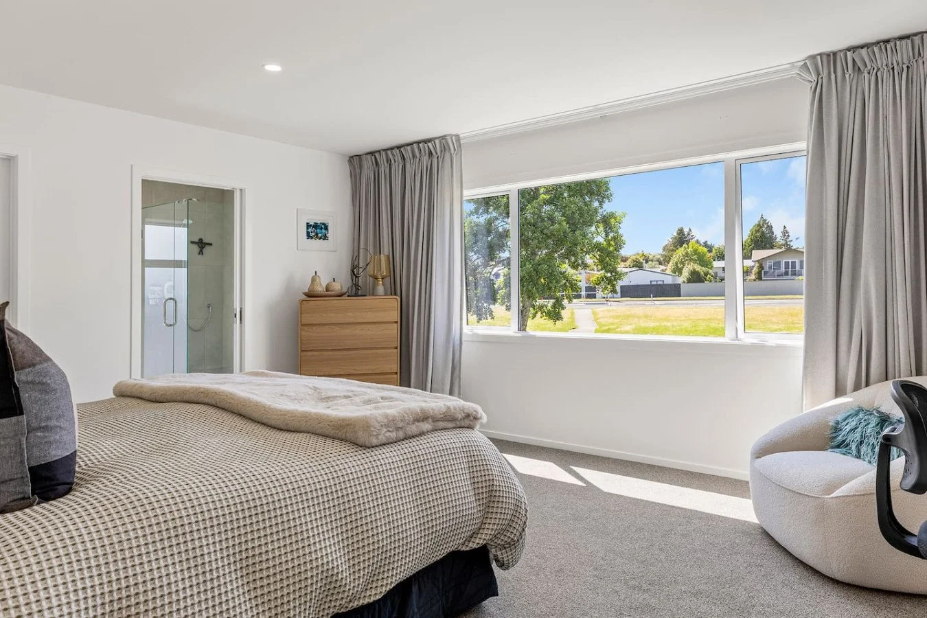 A bedroom with a large window overlooking a lawn and trees, a bed with a checked duvet and a faux fur throw, a white modern chair with pillows, a wooden dresser with decorative items, and a glass-enclosed shower area in the corner.