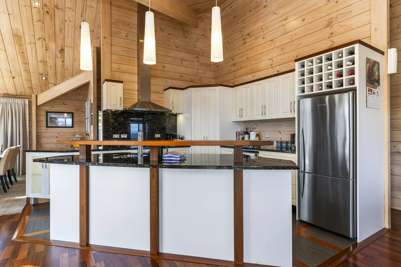 Modern kitchen with white cabinets, black granite countertops, stainless steel refrigerator, wood accents, and pendant lighting, in a log-style home interior.