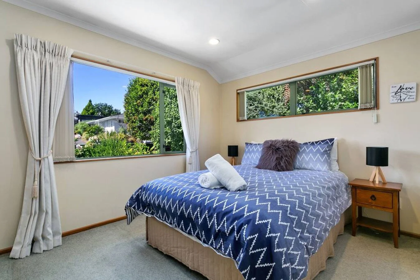A neatly made bed with a blue and white zigzag patterned comforter, a rolled towel, and a fluffy pillow in a bedroom with two large windows and greenery outside.