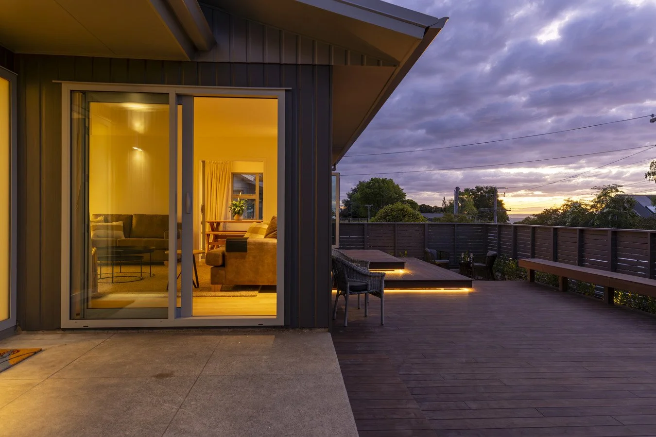 View of a modern house's outdoor wooden deck at sunset with interior lights glowing, visible through glass sliding doors, and a cloudy sky in the background.