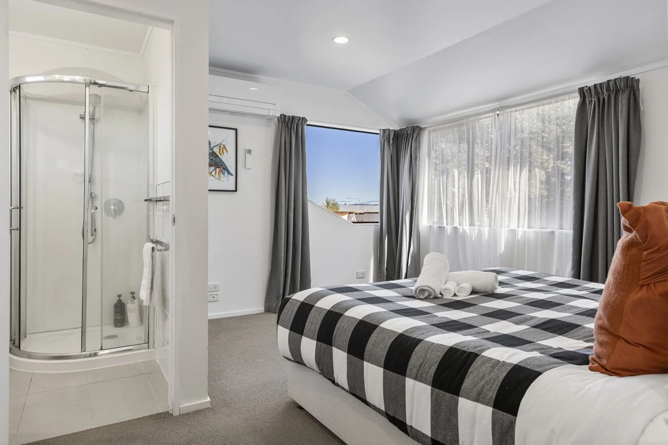 A bedroom with a bed covered in a black and white checkered blanket, decorative pillows, and towels, next to a large window with sheer curtains, and a bathroom with a glass shower enclosure visible to the left.