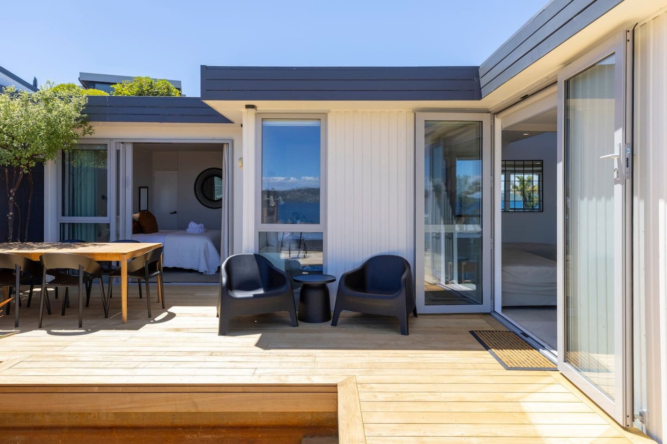 Outdoor balcony with wooden flooring, black chairs, a small round table, and a sliding glass door leading to a bedroom; part of a modern house with glass windows and a view of water and sky.