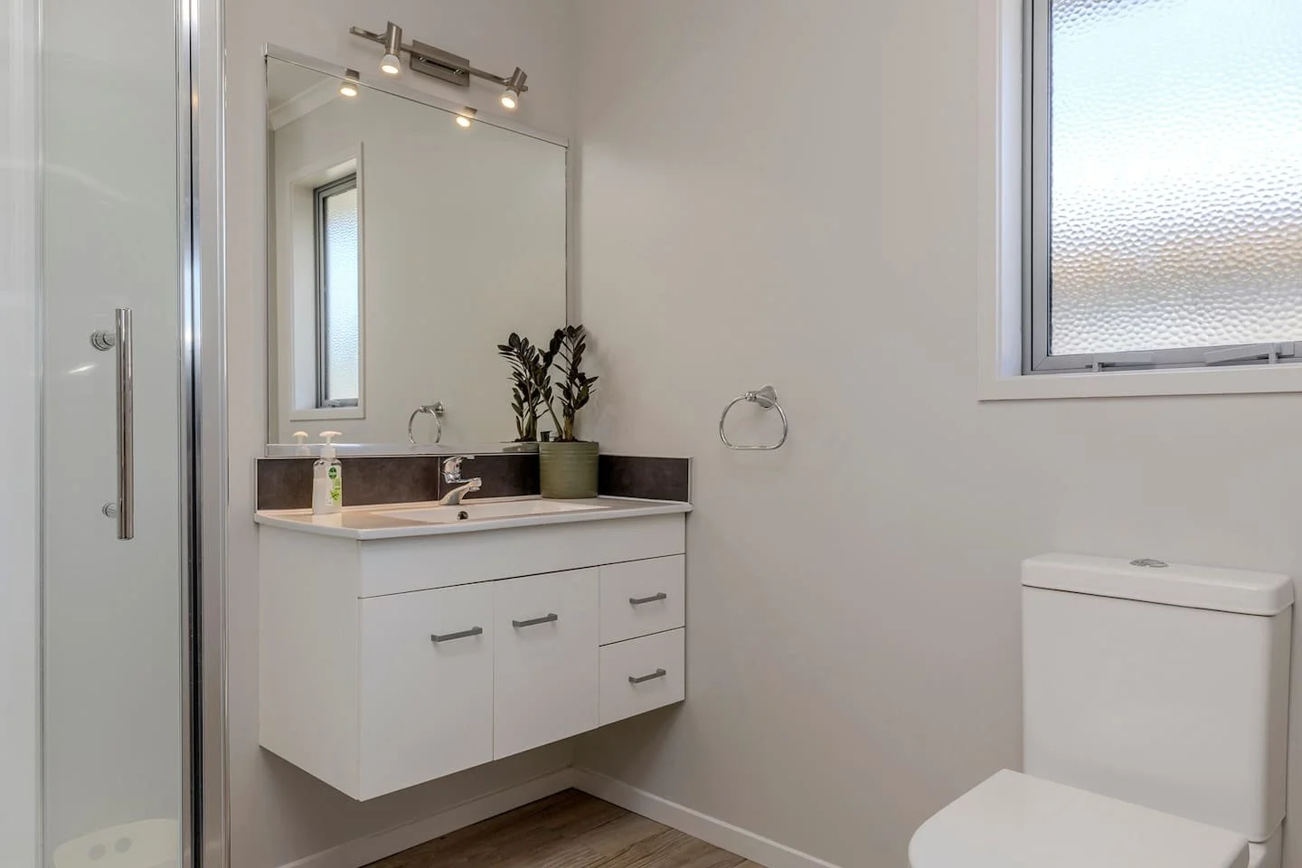 A bathroom with a white vanity, a large mirror, a potted plant, a soap dispenser, a frosted window, and a toilet.