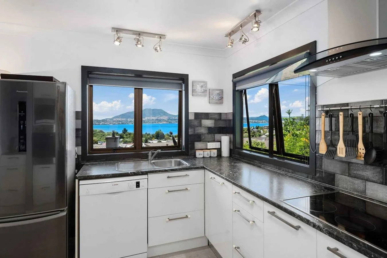 Modern kitchen with white cabinets, black countertop, and large windows showing a scenic view of water, trees, and mountains.