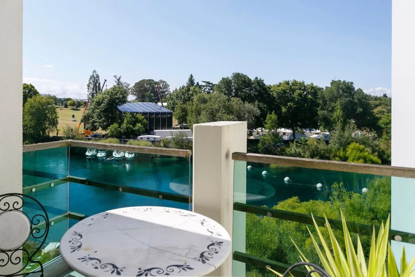 A balcony with a small round table and a black wrought iron chair, overlooking a swimming pool surrounded by greenery, trees, and a large outdoor stage with a blue canopy in the distance on a sunny day.