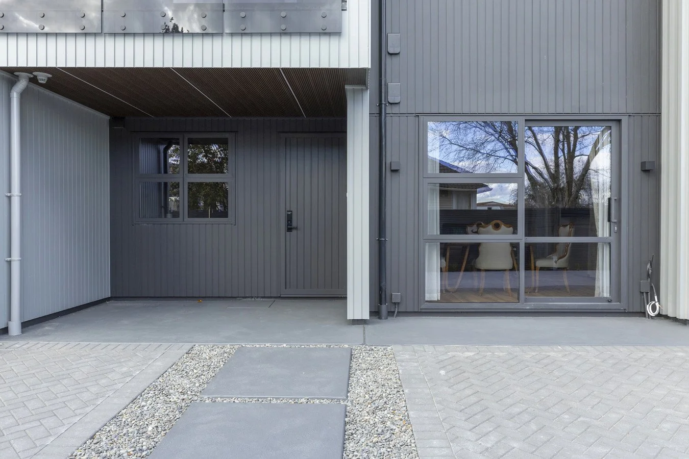Exterior view of a modern residential building with a gray panel facade, featuring a small window, a door with an intercom, and a large sliding glass door revealing a dining area with chairs inside, with a tree reflected in the glass.