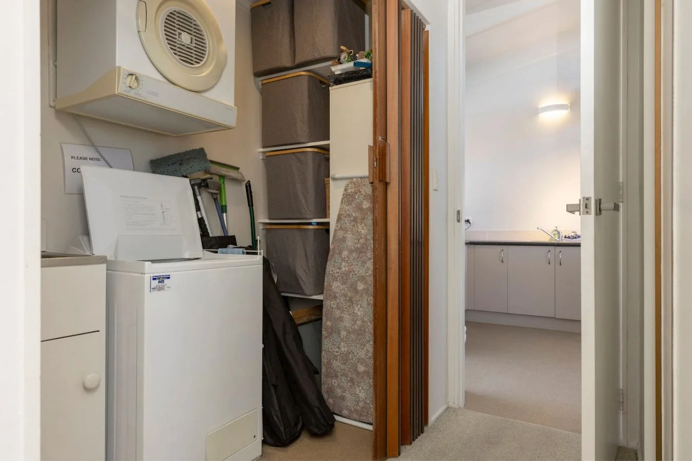 Laundry room with washing machine, laundry supplies, and storage bins; doorway leads to kitchen with sink and cabinets.