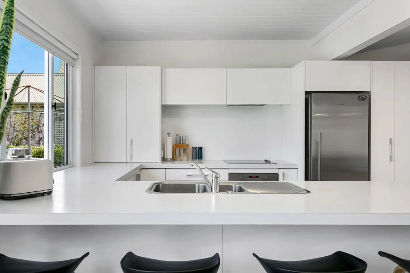 Modern white kitchen with a window, stainless steel refrigerator, sink, and black chairs in the foreground.