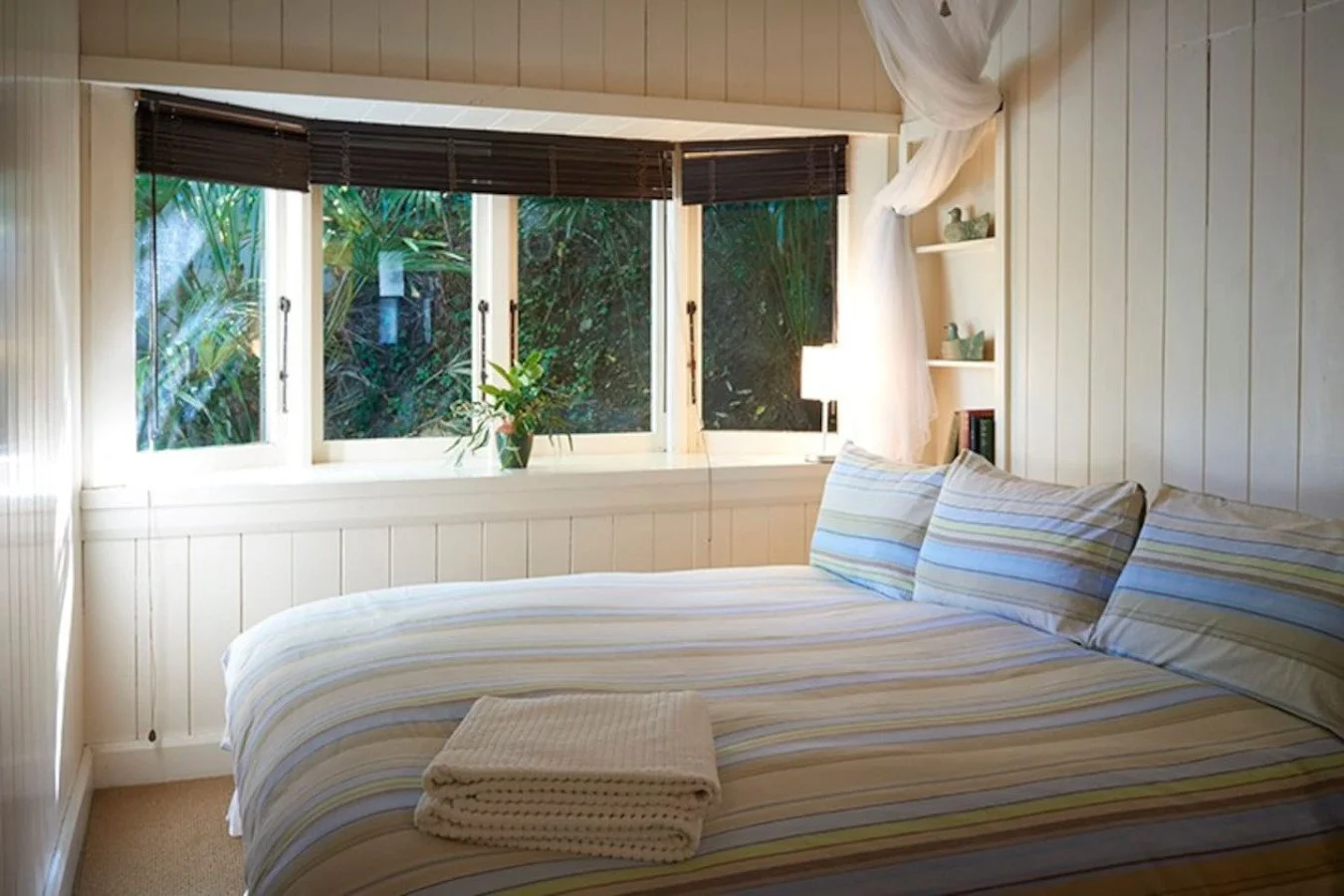 A cozy bedroom with a bed covered in striped bedding, a window with a view of lush greenery, and a small shelf with books and decorative items.