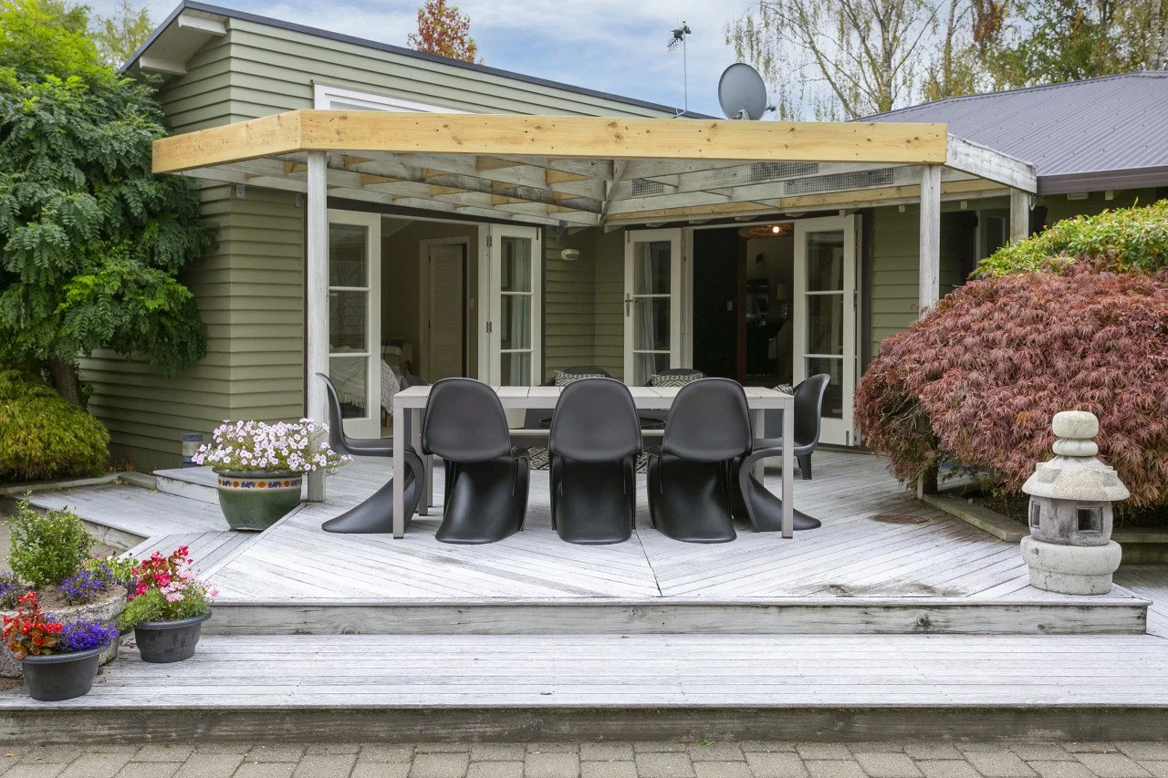 Backyard patio with a wooden deck, outdoor dining table with black chairs, potted flowers, lush bushes, and a green house with French doors.