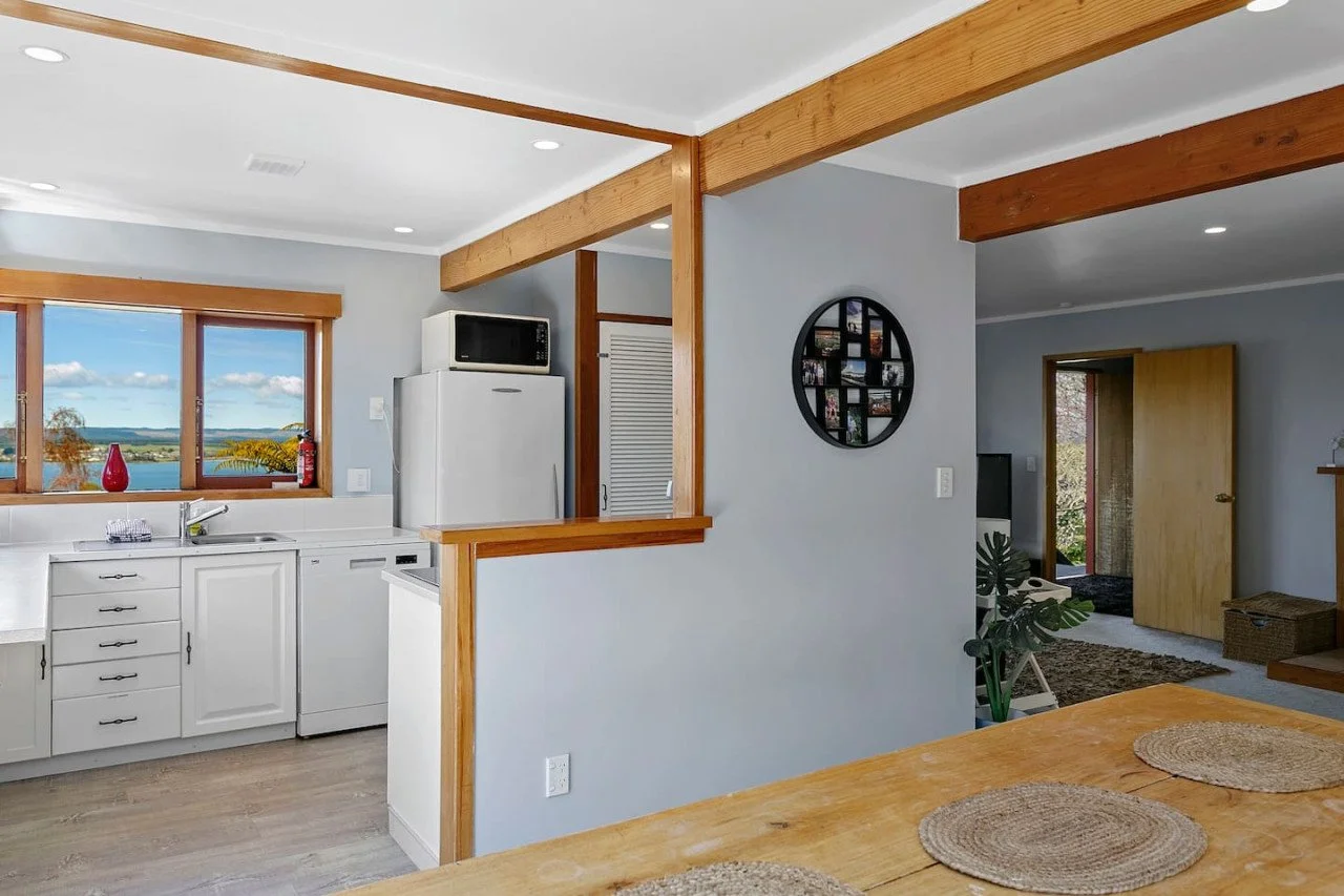 A cozy kitchen with white cabinets, a large window overlooking a lake, and a microwave on top of a refrigerator. The room features wooden accents and a view into a living area with a door and a small outdoor space.