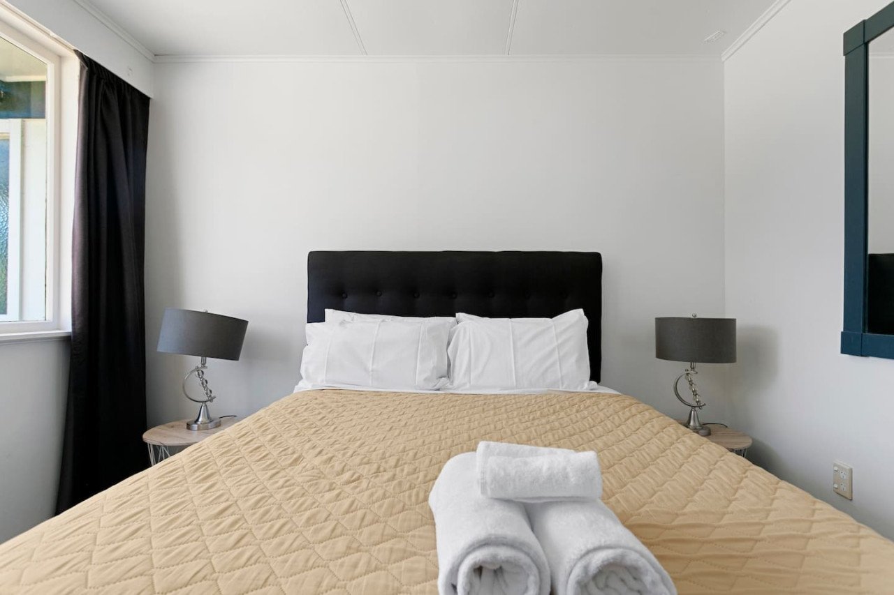 A neatly made bed with white pillows and a beige quilted blanket, with three rolled white towels on top, in a bedroom with two gray bedside lamps and a window with dark curtains.