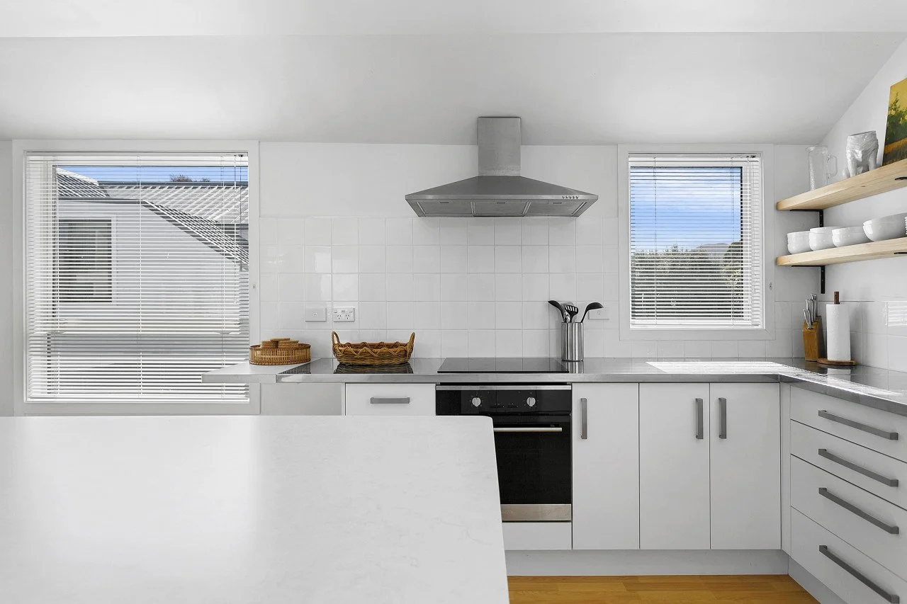 A bright, white kitchen with two windows, white cabinets, a stovetop, a stainless steel range hood, and open wooden shelves with white bowls and mugs.