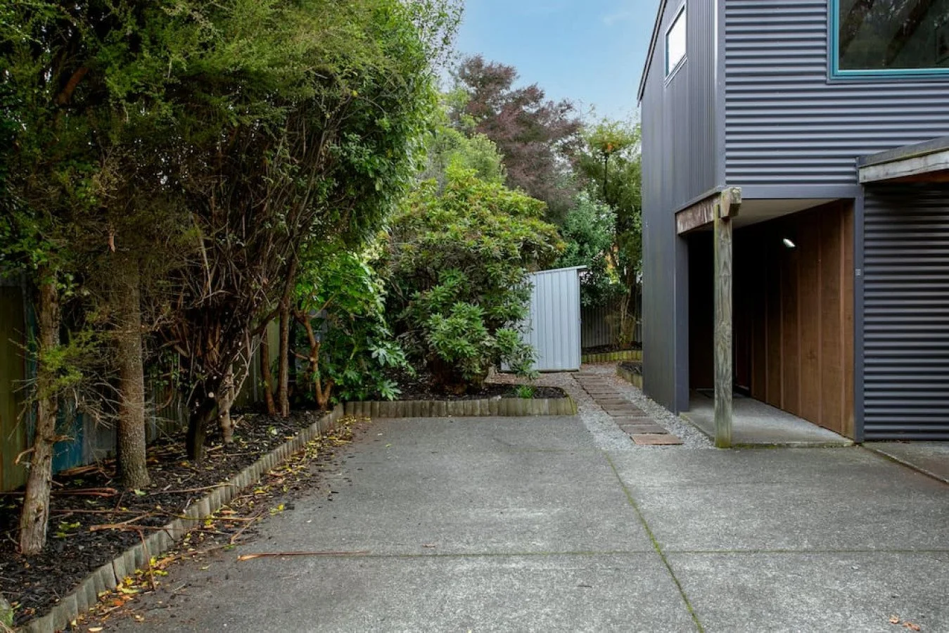 A small paved courtyard area next to a modern gray building with wooden accents, surrounded by lush green trees and bushes, with a small gray shed in the background.