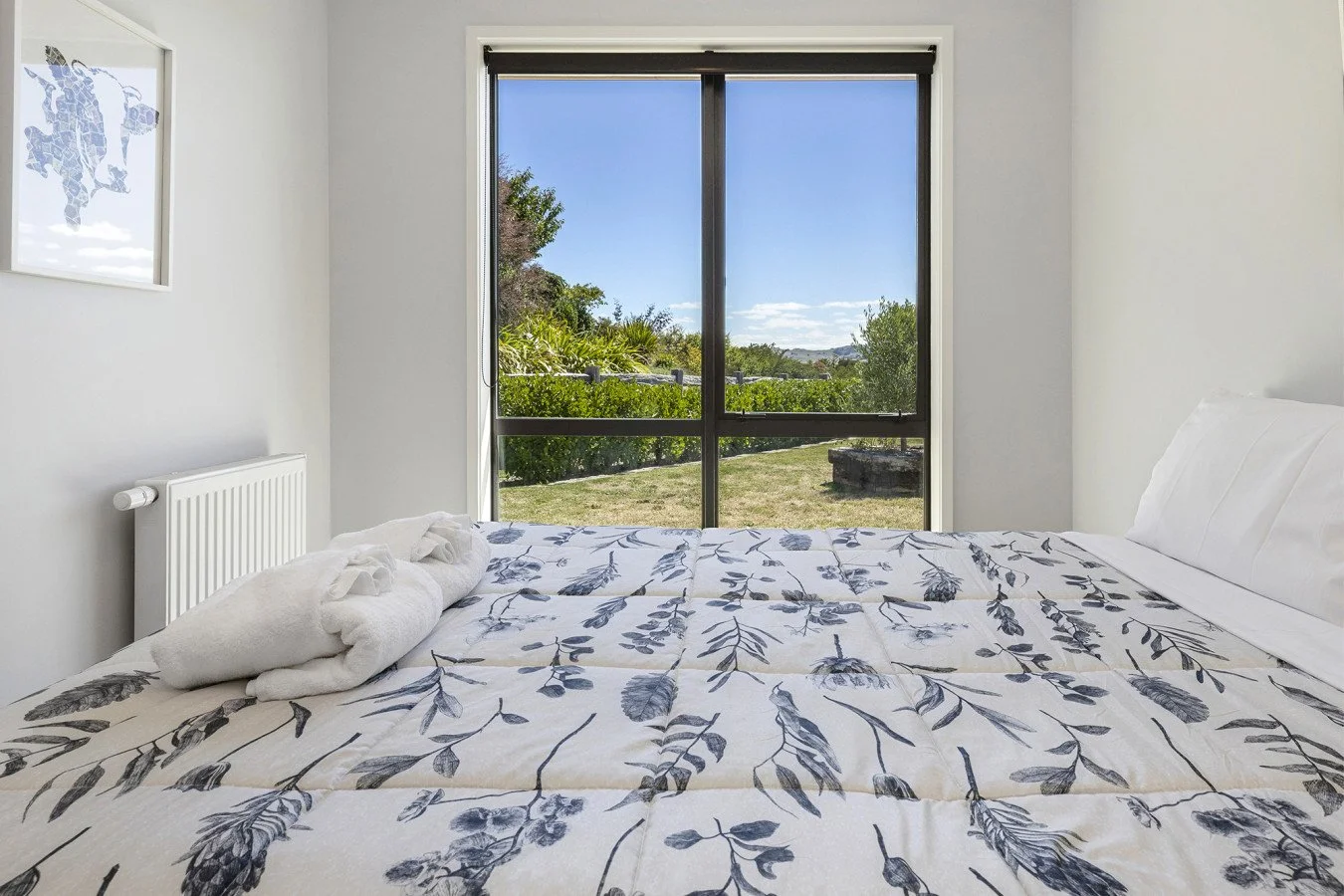 A bedroom with a bed covered in a white and black floral patterned comforter, a rolled towel on top, a white pillow, a window with black blinds showing a sunny outdoor scene with greenery and a clear blue sky, and a wall art piece on the left side.