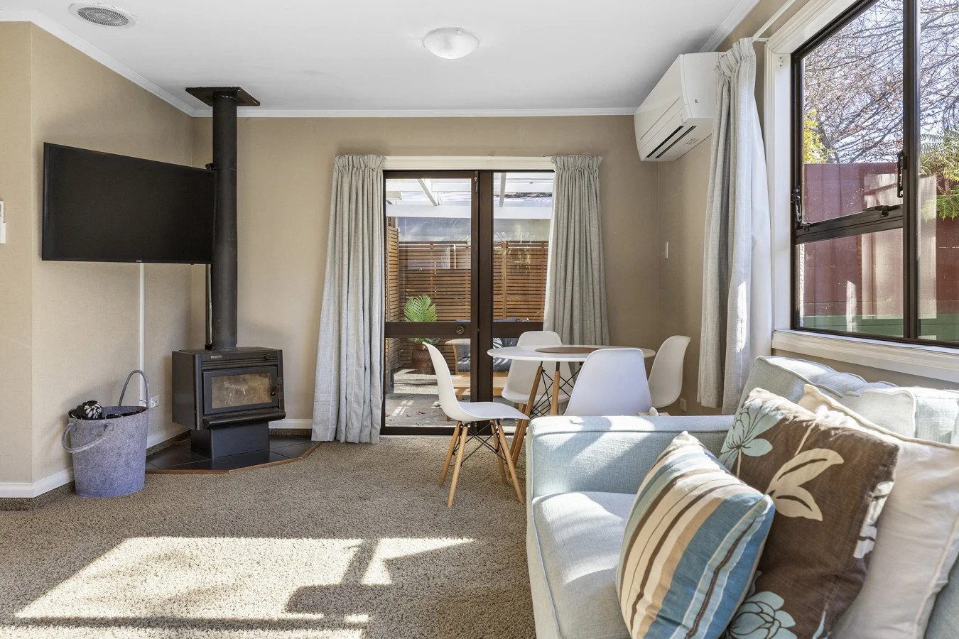 Living room with beige walls, carpeted floor, wall-mounted TV, wood stove, sliding glass door to outdoor patio, round dining table with four white chairs, window with curtains, air conditioning unit, and a light green sofa with patterned pillows.