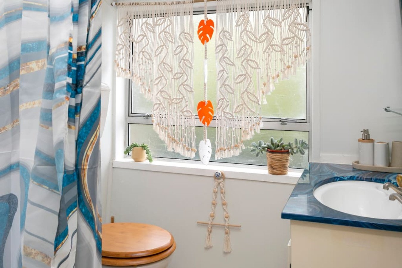 A bathroom with patterned curtains on the window, a small potted plant, and a macramé wall hanging with orange, white, and beige leaves, and a dreamcatcher hanging below.