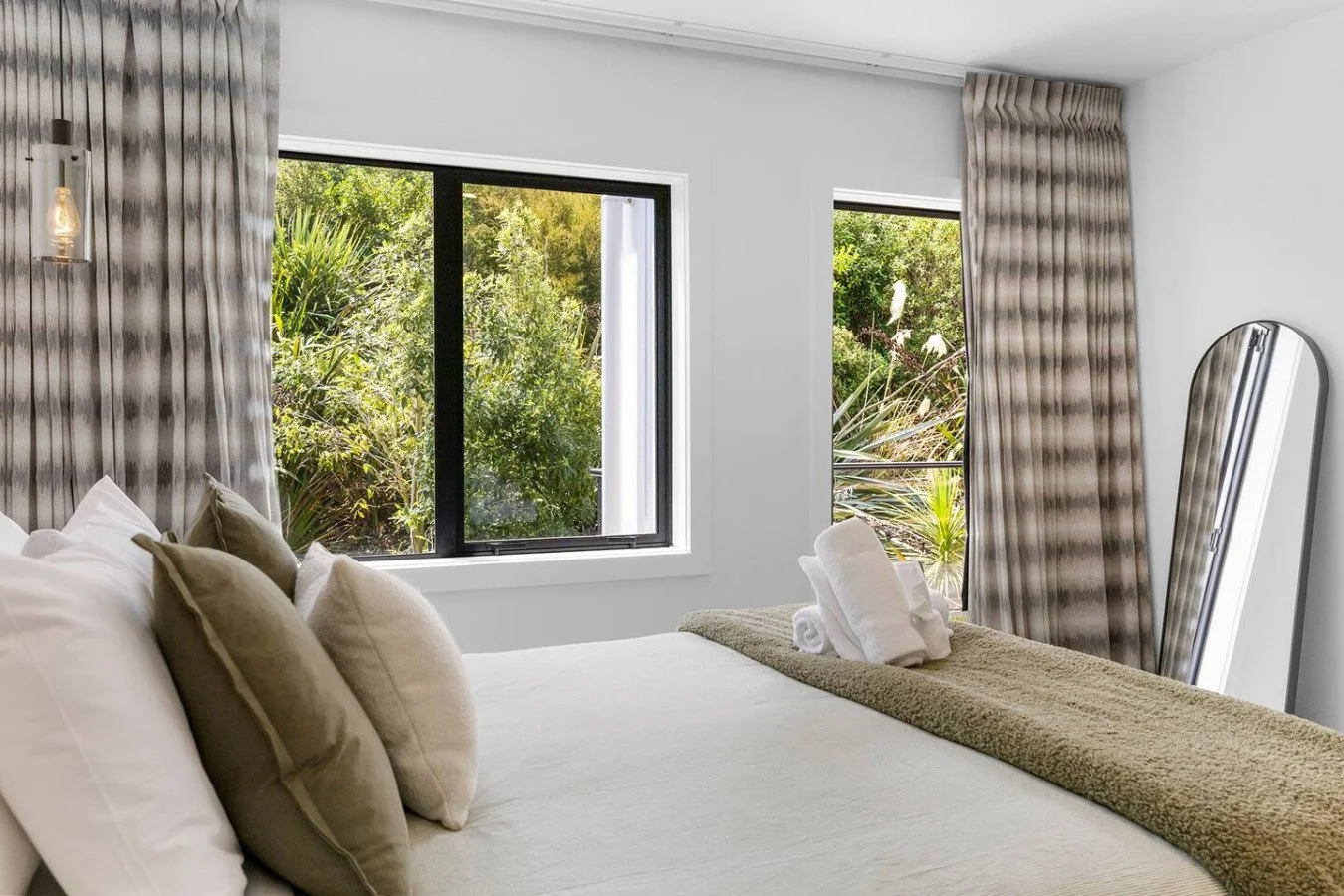Bedroom with white walls, large windows showing greenery outside, beige and white bedding, rolled white towels on bed, striped curtains, and a leaning mirror.