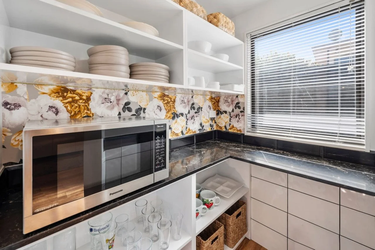 Kitchen corner with open shelves holding white dishes, a microwave, a window with blinds, floral backsplash, and black countertops.