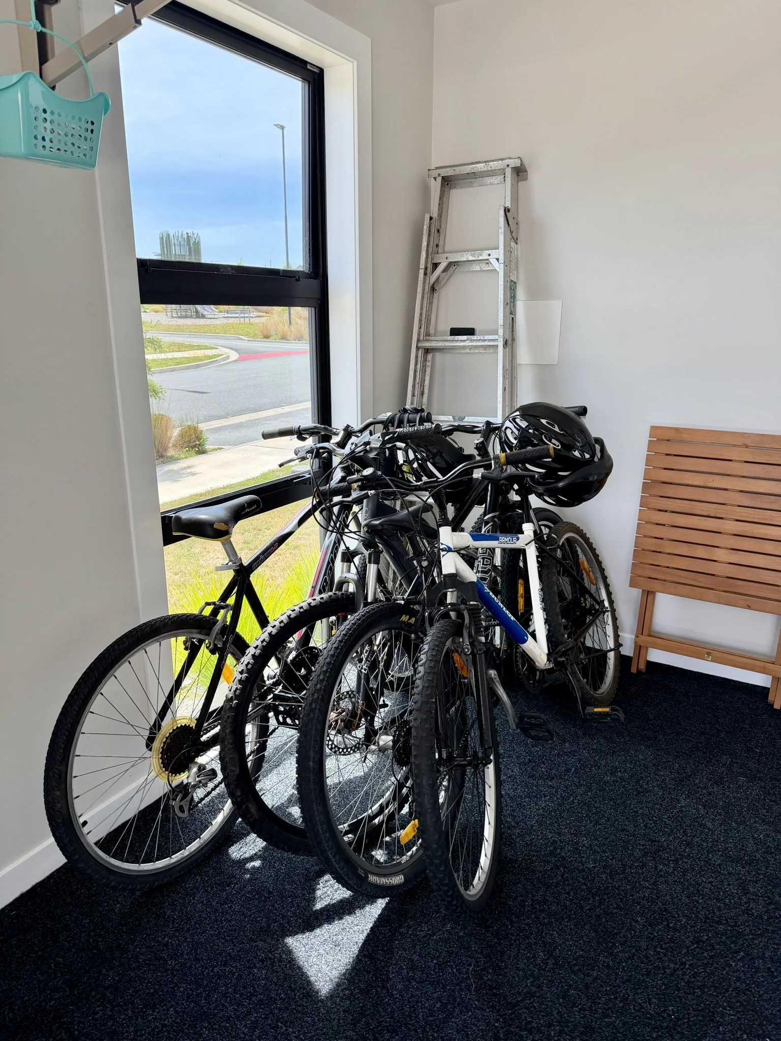 Three bicycles, one with a helmet on the handlebar, leaning against a glass window in a room with white walls, a wooden bench, a ladder, and a turquoise basket hanging on the wall.
