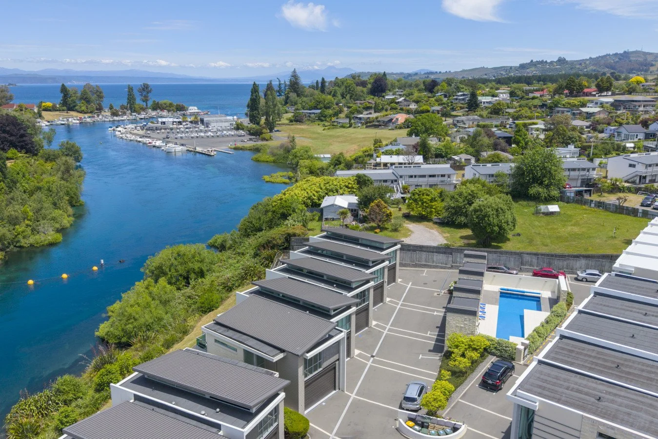 Aerial view of modern townhouses next to a river with a marina and boats, surrounded by lush greenery and rolling hills under a clear blue sky.