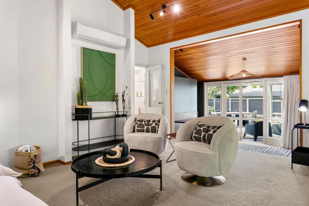 Living room with white walls, tan carpet, and a wooden ceiling. It has two white armchairs with patterned pillows, a black coffee table with decorative objects, a black side table with a lamp, and a green abstract wall art piece. A sliding glass door