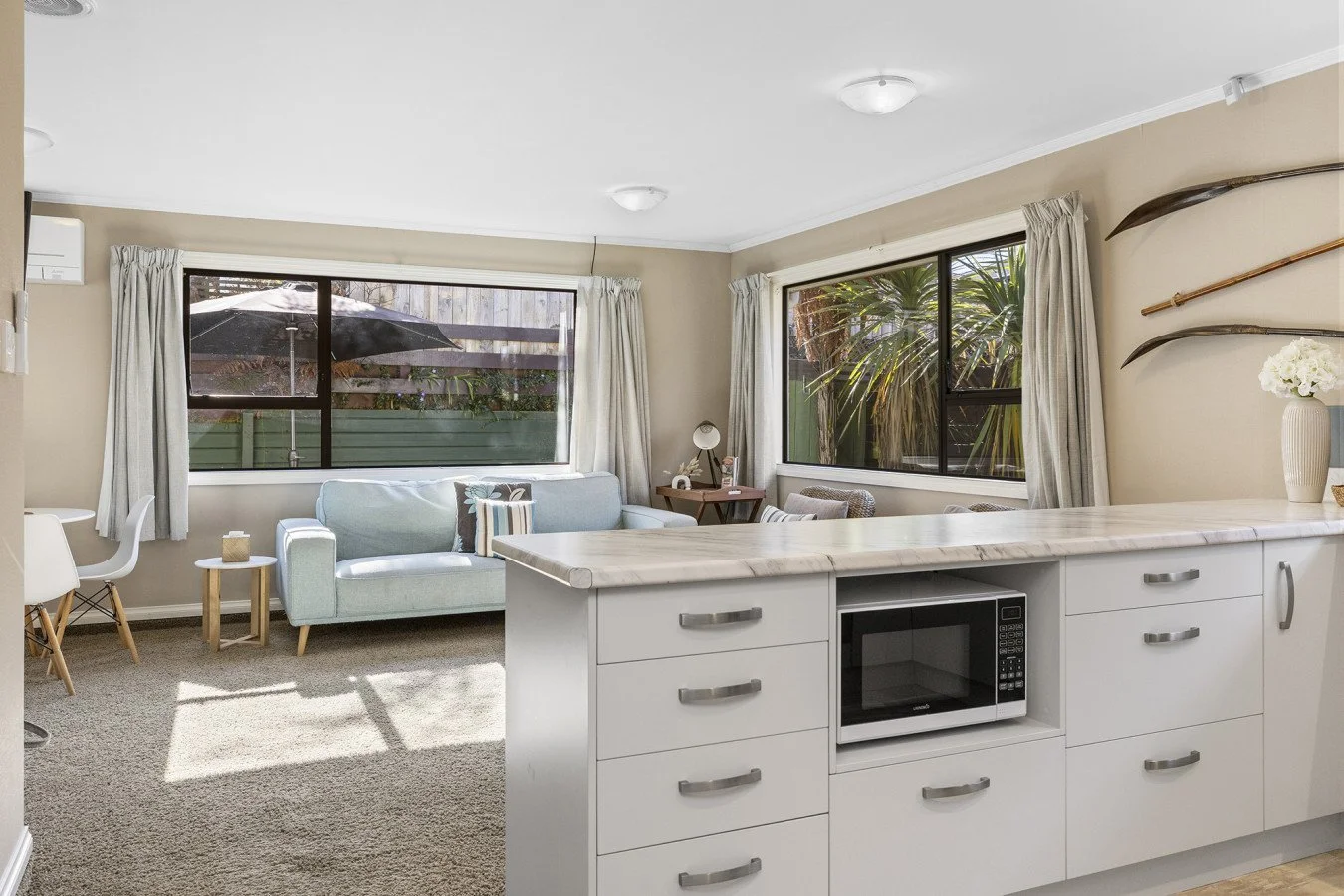 Living room with large windows, sofa, small side tables, a desk with a chair, and decorative wall accents, viewed from the kitchen area with white cabinets and a microwave.