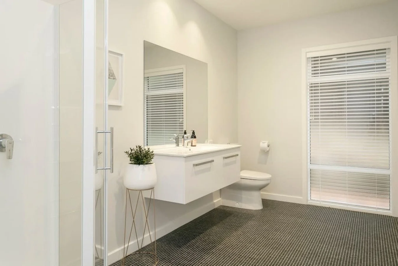 Modern bathroom with white vanity, large mirror, toilet, window with blinds, and patterned gray carpet.