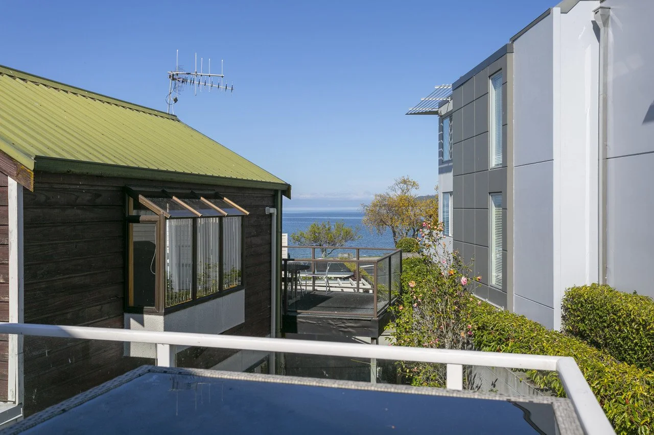 View of residential buildings with a balcony overlooking the ocean, trees, and a clear blue sky.