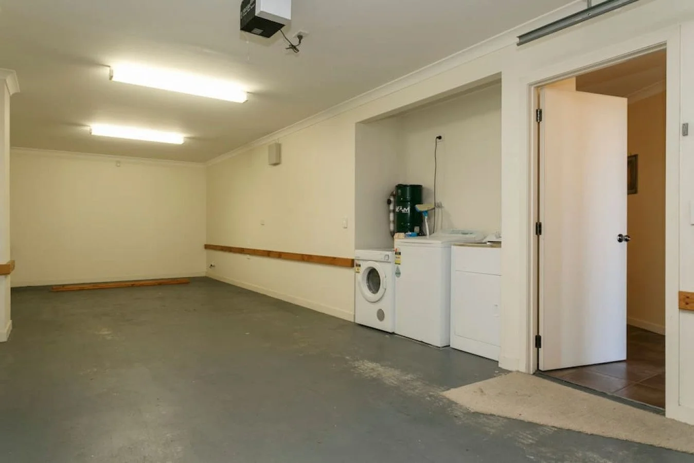 Empty room with laundry area containing a washing machine, a dryer, and water filtration system. The room has beige walls, fluorescent lighting, and a sliding door leading to another room with brown tile flooring.