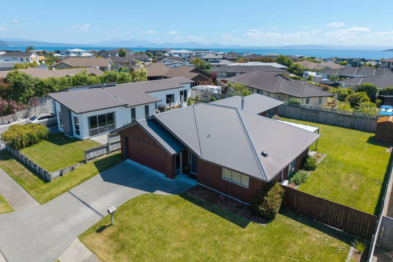 Aerial view of a suburban neighborhood with houses, green lawns, and a distant ocean in the background, under a partly cloudy sky.