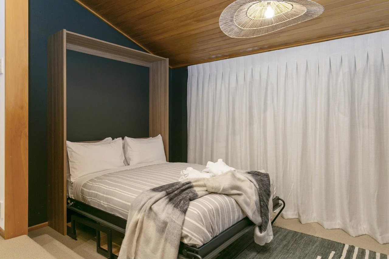A cozy bedroom with a bed that has white linens and a gray striped blanket, situated in front of white curtains and illuminated ceiling light.