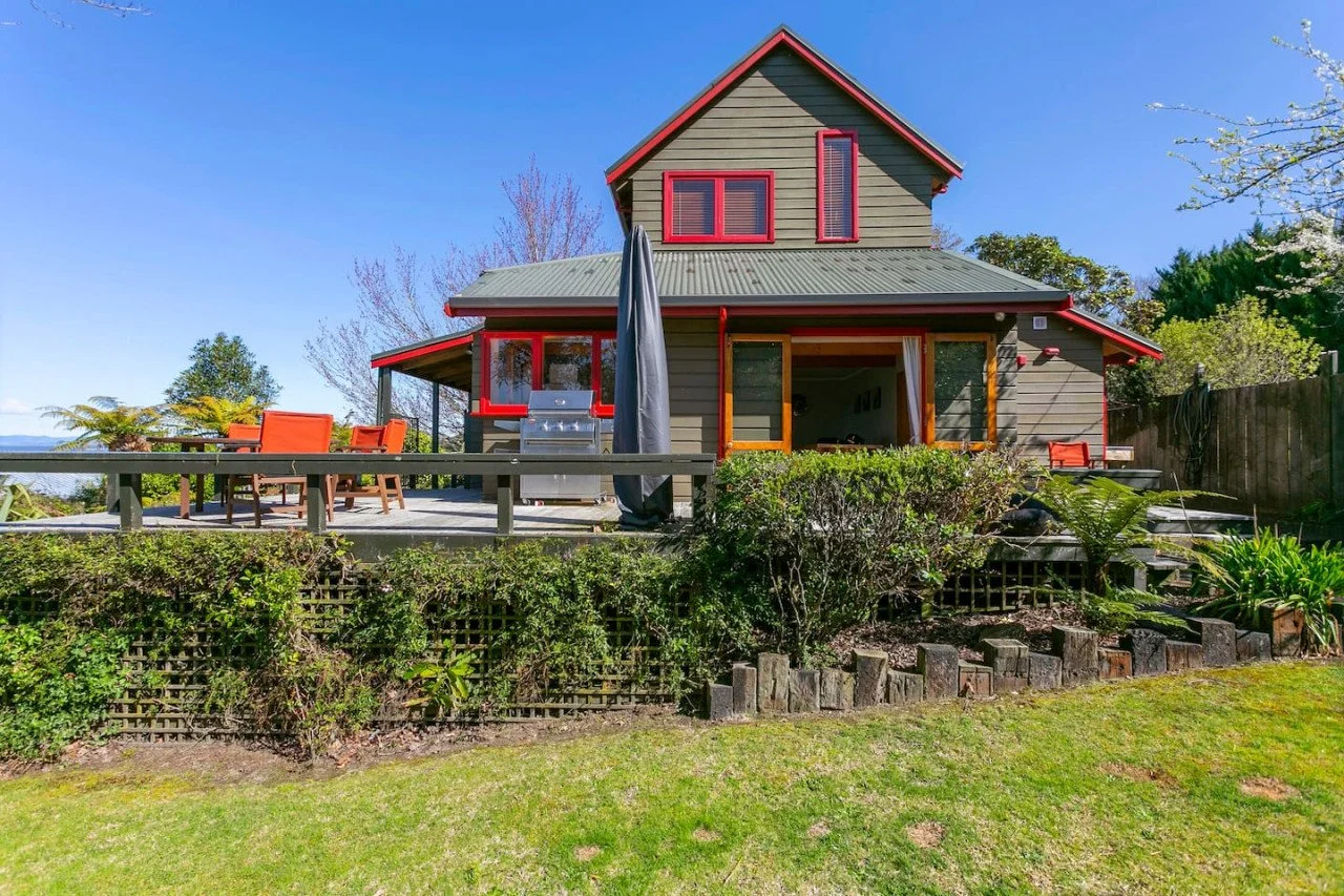 A two-story house with a gray exterior and red trim, a green metal roof, and a deck with outdoor furniture and a grill, set in a lush green yard under a clear blue sky.